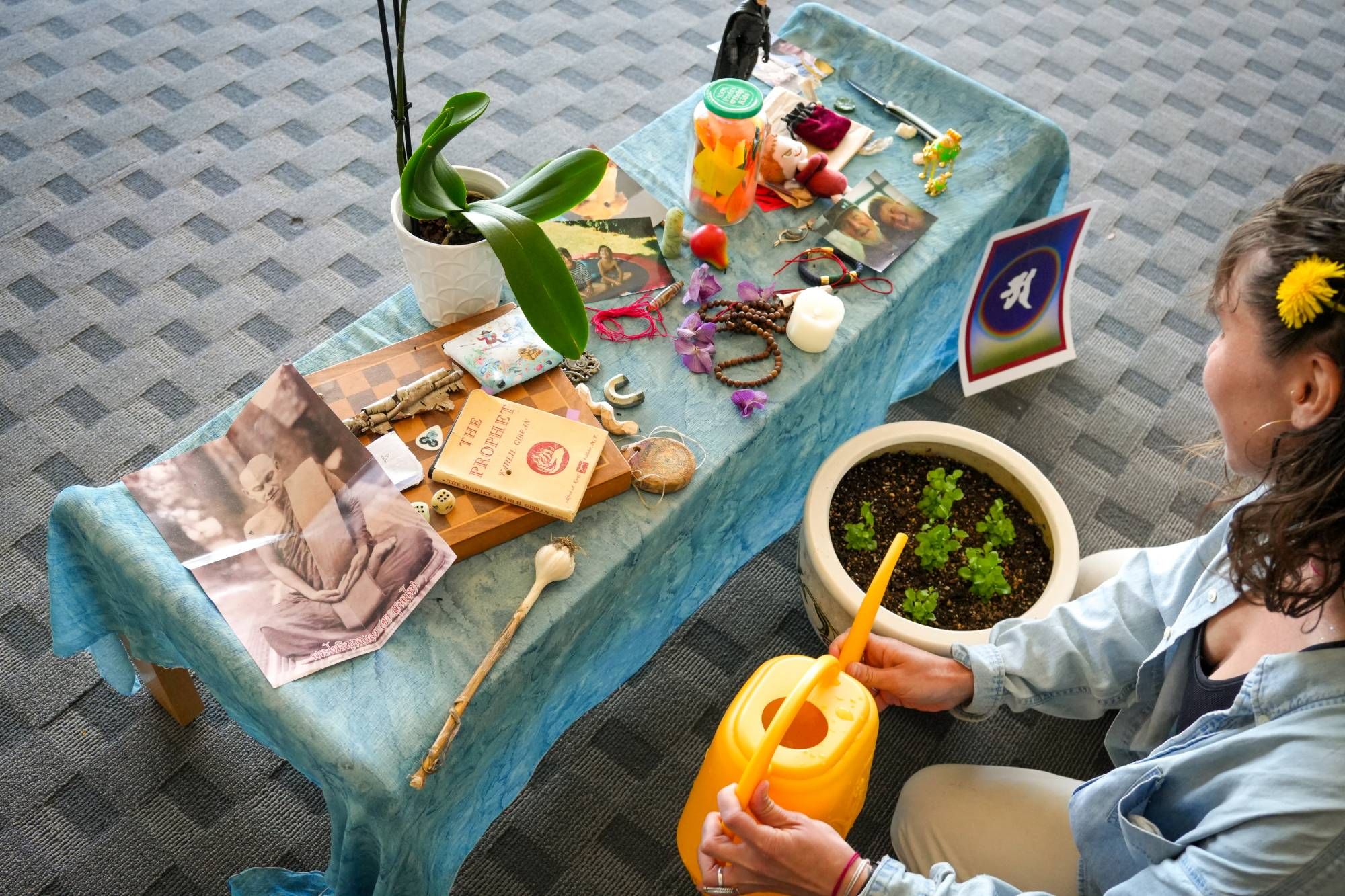 A person with a dandelion in their hair waters a small potted plant beside a low altar of photographs, books, beads and flowers on a blue cloth.