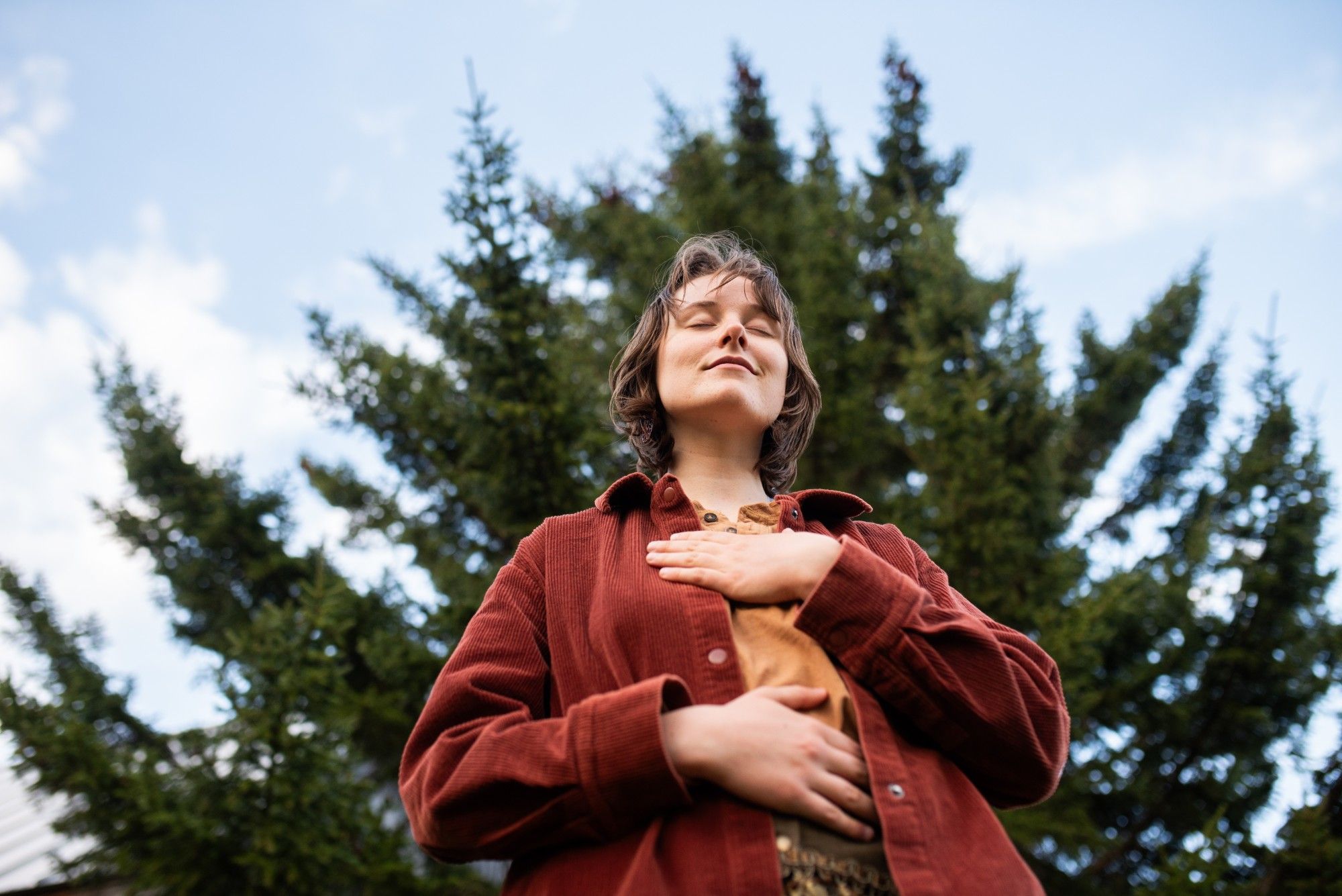 A person with eyes closed and a hand on their chest and belly, standing beneath a tall evergreen against a blue sky.