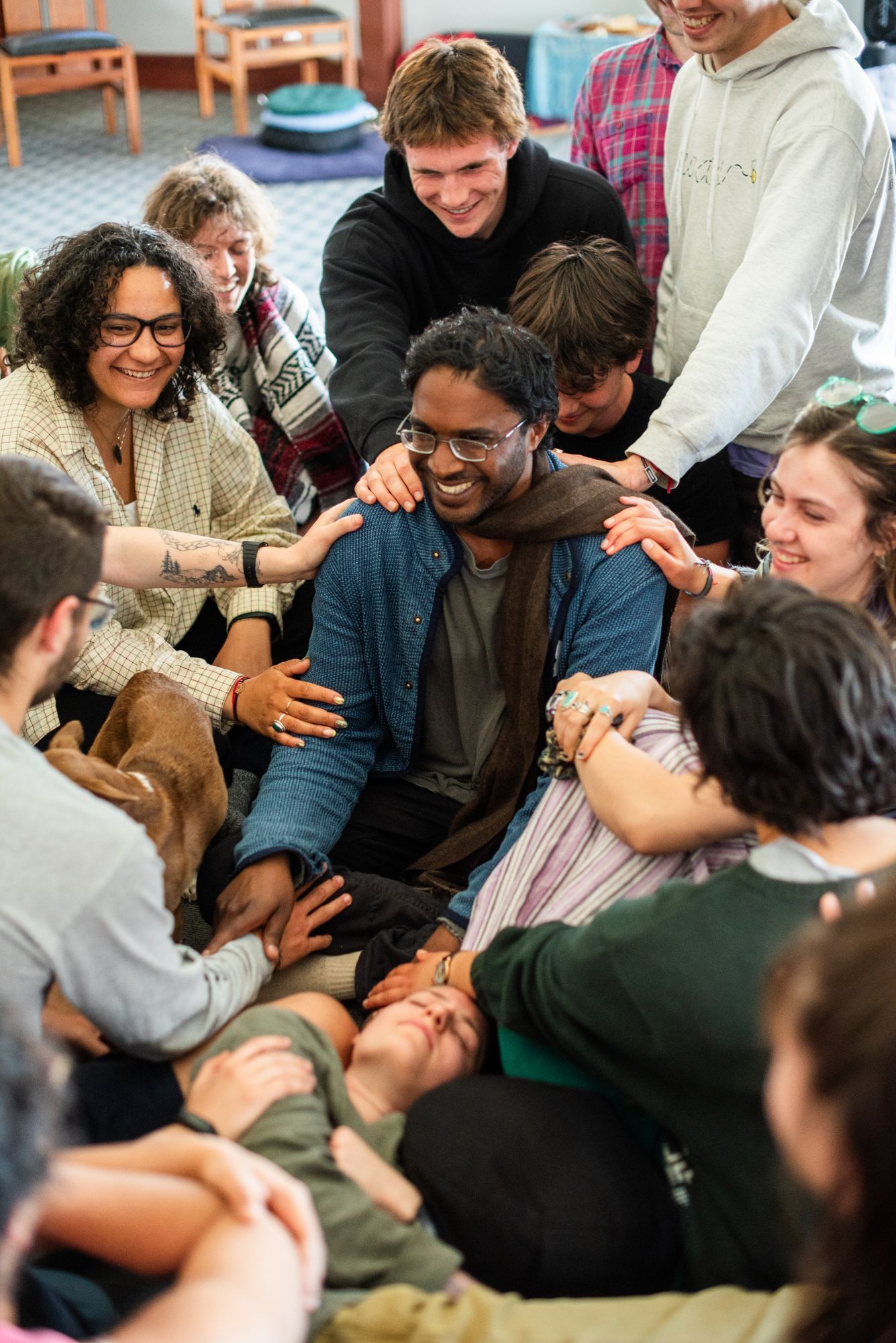 A circle of young people sitting close together, laughing, with hands resting on the shoulders of the man at the center.