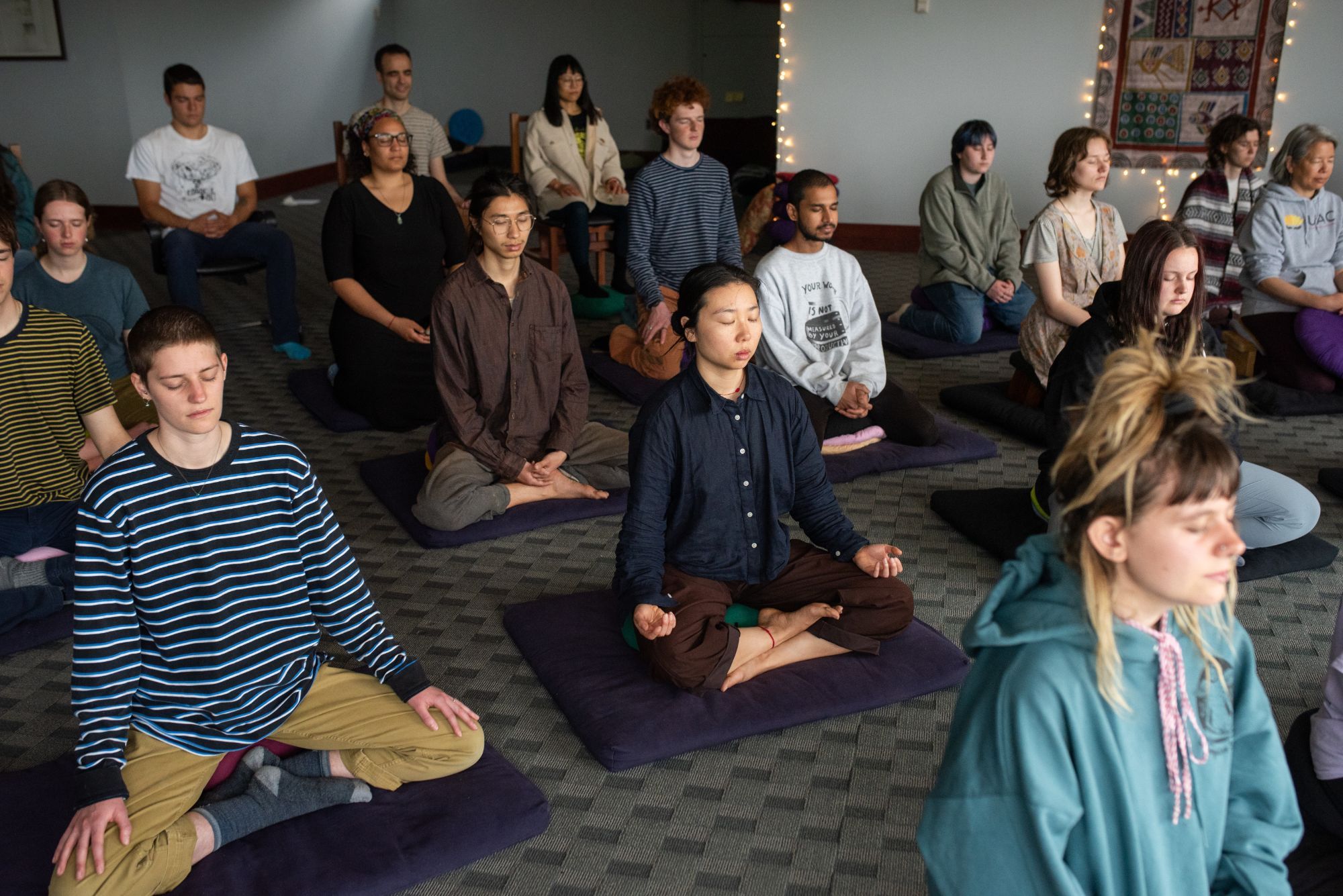 A room of people seated in meditation on dark cushions, eyes closed, with string lights and a woven wall hanging behind them.