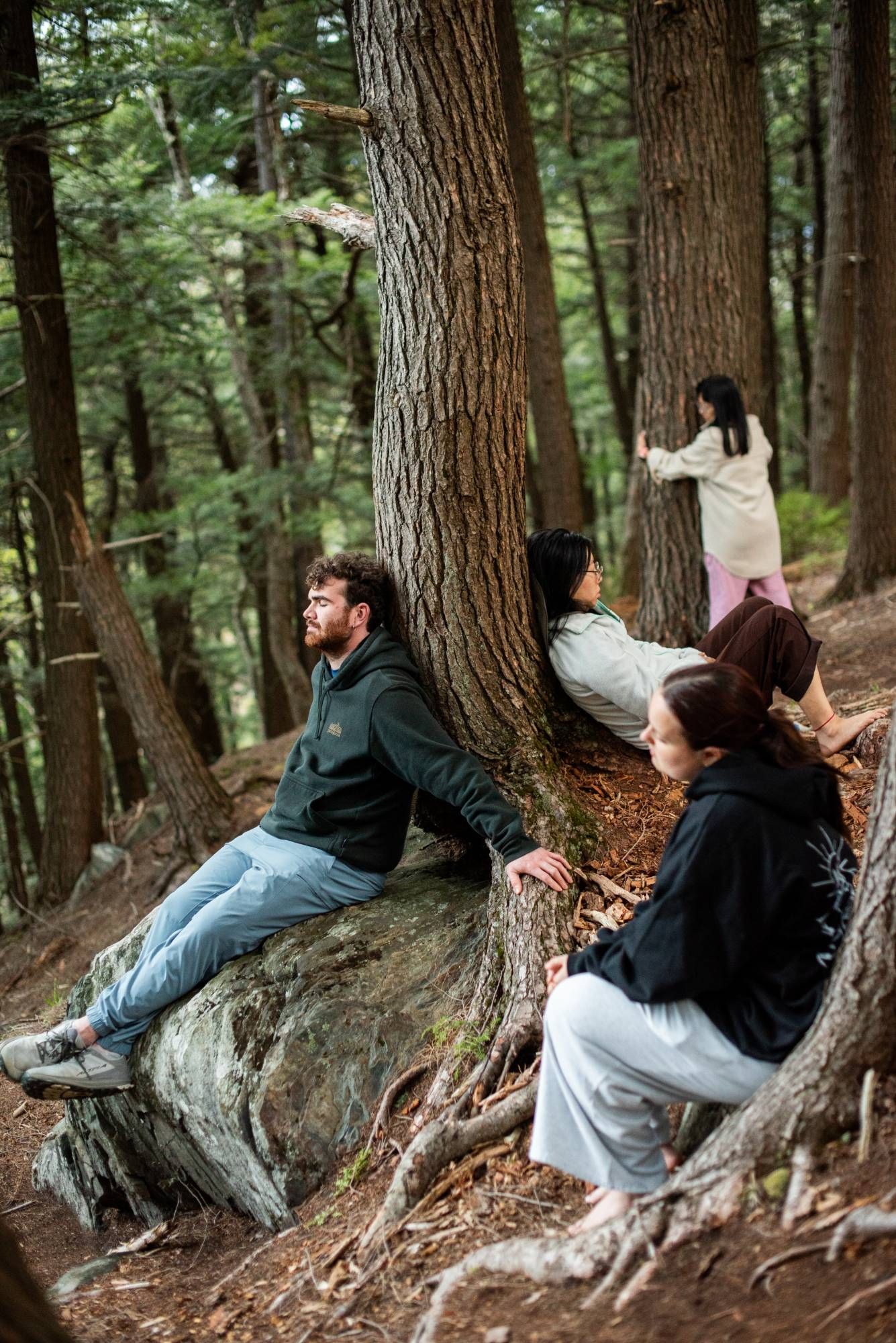 People resting against trees and a boulder in a forest, some with eyes closed.
